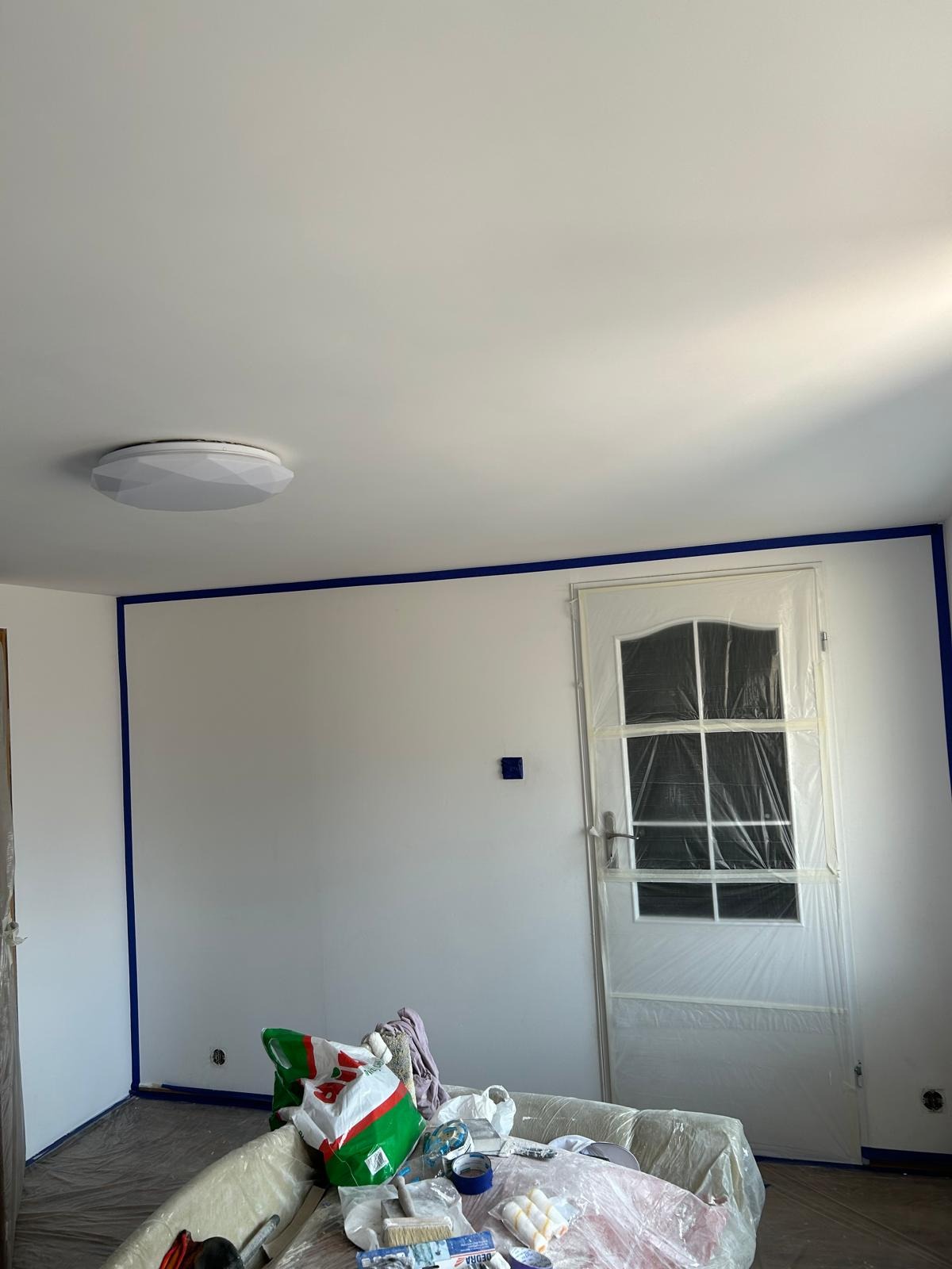 Przygotowane do malowania wnętrze pokoju z zabezpieczonymi listwami przypodłogowymi i framugą drzwi taśmą malarską oraz folią ochronną, widoczne narzędzia malarskie i wiadro z farbą.