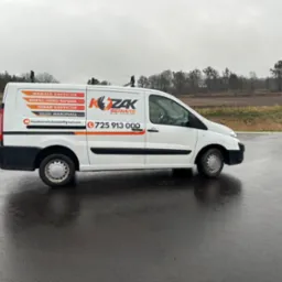 Biały samochód dostawczy firmy elektrycznej 'KZAK Serwis' z widocznym logo i numerem telefonu, zaparkowany na mokrym asfalcie w pochmurny dzień.