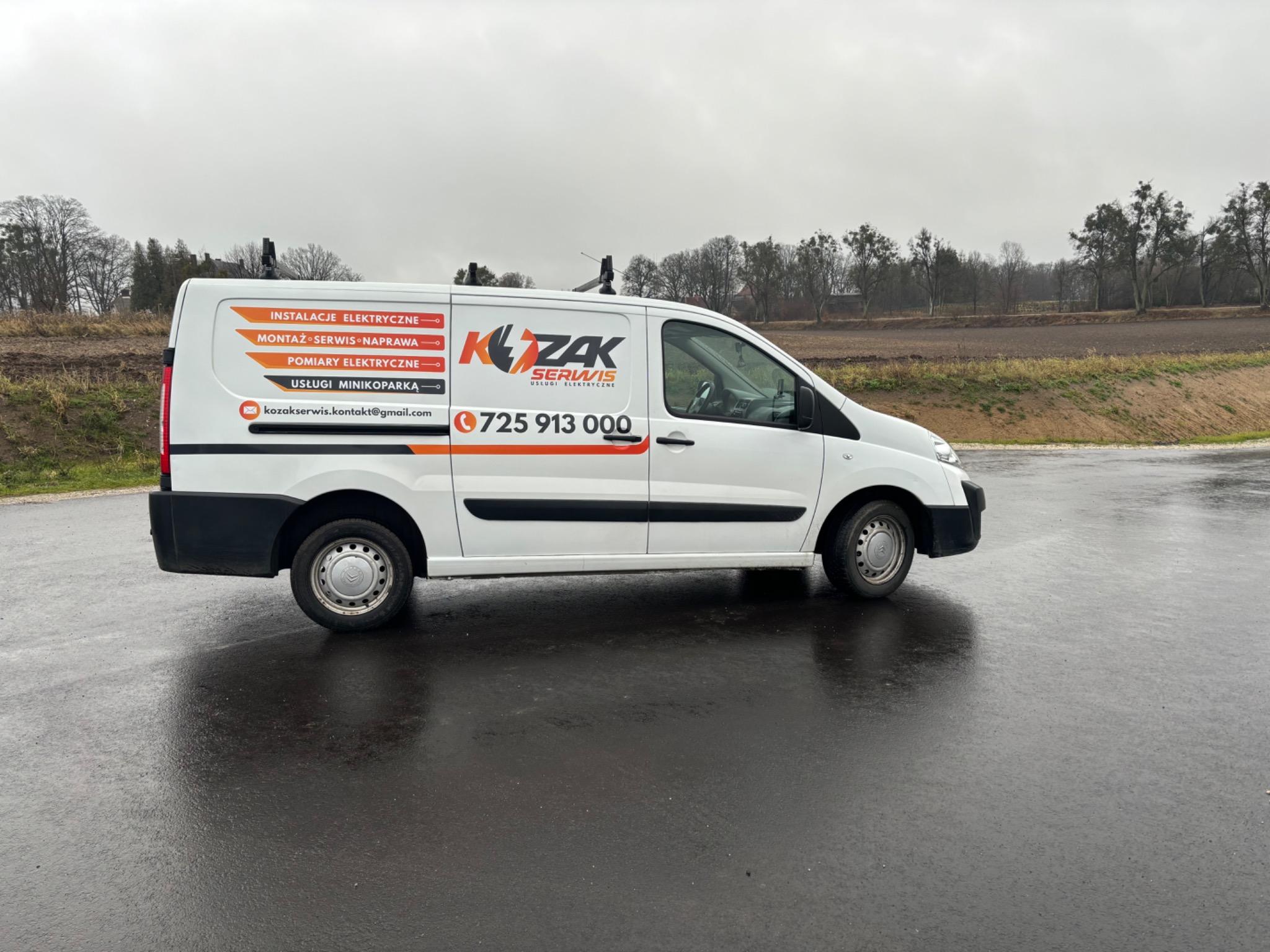 Biały samochód dostawczy firmy elektrycznej 'KZAK Serwis' z widocznym logo i numerem telefonu, zaparkowany na mokrym asfalcie w pochmurny dzień.