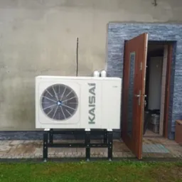 Biała pompa ciepła marki Kaisai zamontowana na czarnej, metalowej konstrukcji przed wejściem do budynku z otwartymi drzwiami. Widoczny fragment trawnika.