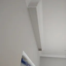 Kątny widok na białą sztukaterię sufitową, zamontowaną w rogu pomieszczenia, z widocznym fragmentem okna zabezpieczonego taśmą malarską.