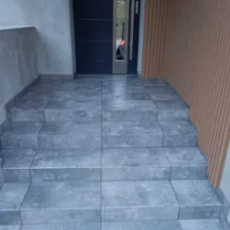 Schody wejściowe wyłożone szarymi płytkami imitującymi kamień, prowadzące do drzwi wejściowych z elementami dekoracyjnymi, z widocznymi surowymi ścianami po bokach i drewnianą okładziną ścienną po...