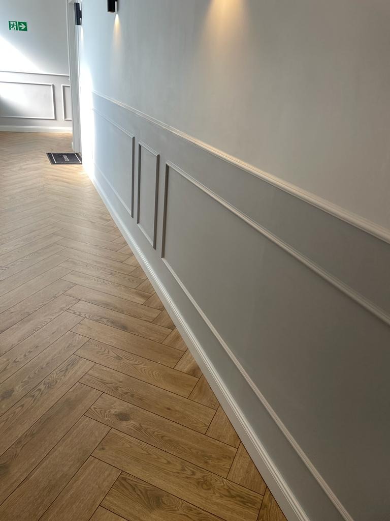 Korytarz z drewnianą podłogą ułożoną w jodełkę i szarą ścianą z dekoracyjnymi panelami, oświetlony punktowymi lampami sufitowymi.