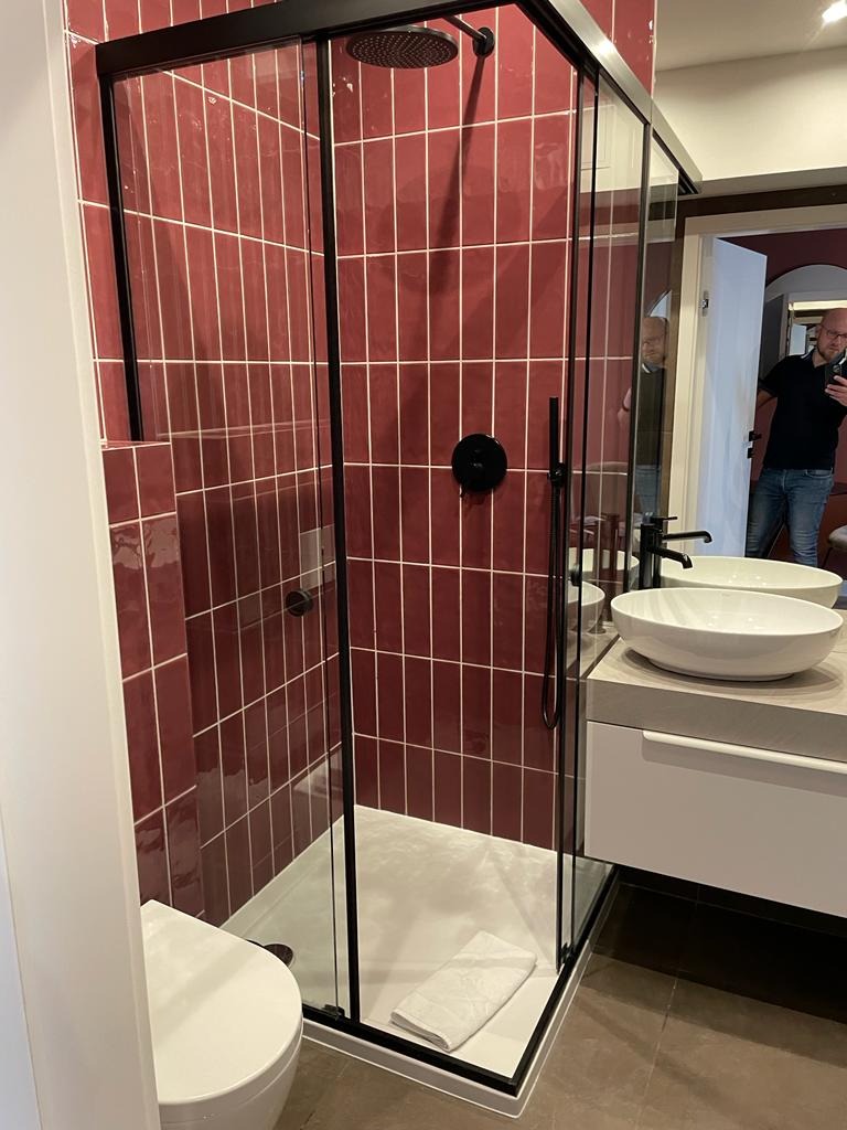 Nowoczesna łazienka z kabiną prysznicową typu walk-in, wyłożona bordowymi płytkami w układzie pionowym z jasną fugą, czarna armatura, biała umywalka nablatowa i toaleta.