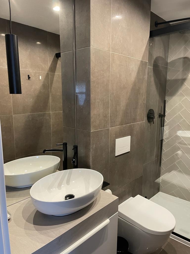 Nowoczesna łazienka z szarymi płytkami, dwiema owalnymi umywalkami nablatowymi, czarną armaturą, wiszącą lampą i kabiną prysznicową z płytkami ułożonymi w jodełkę.