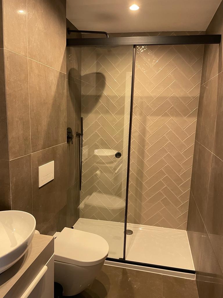 Nowoczesna łazienka z kabiną prysznicową typu walk-in z czarnymi profilami, beżowymi płytkami ułożonymi w jodełkę i wiszącą miską WC.