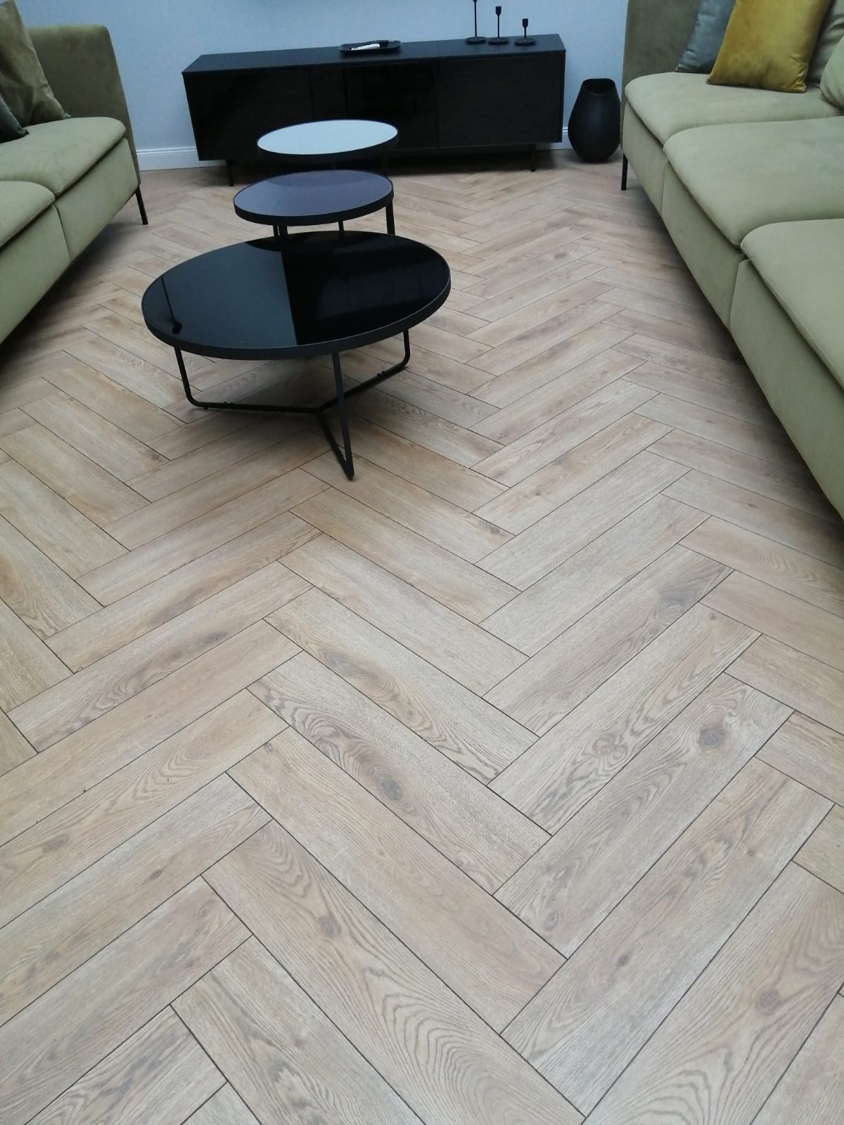 Jasne panele ułożone we wzór jodełki francuskiej w salonie z oliwkowymi sofami i czarnym stolikiem kawowym.