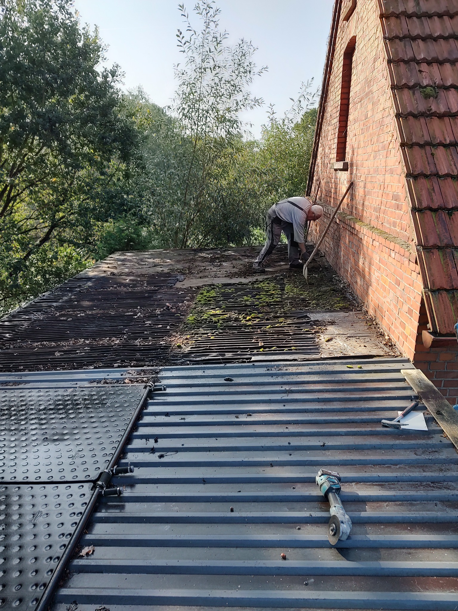 Pracownik usuwa mech i zanieczyszczenia z dachu budynku, widoczne narzędzia i fragment nowej blachy na dachu.