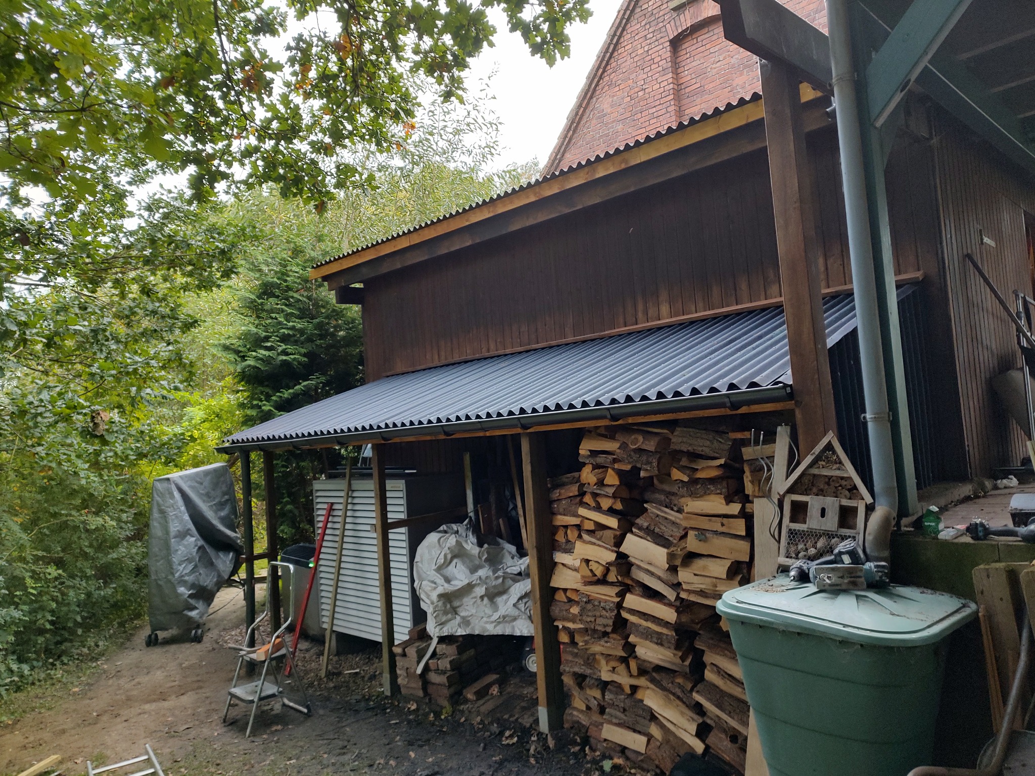 Zadaszona wiata z nowym, szarym, falistym pokryciem dachowym, obok sterta drewna kominkowego, plastikowy pojemnik na deszczówkę i domek dla owadów.