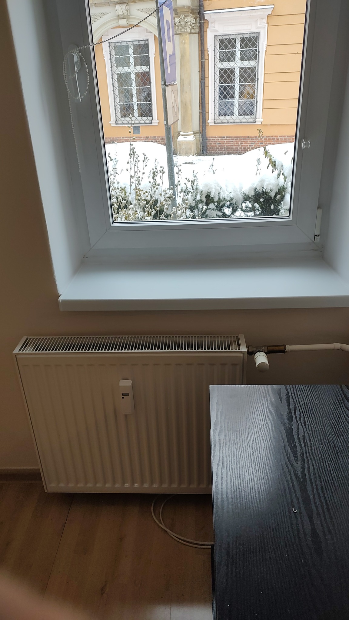 Biały grzejnik panelowy pod oknem na poddaszu w Opolu, widok na zaśnieżony ogród i zabytkową architekturę.