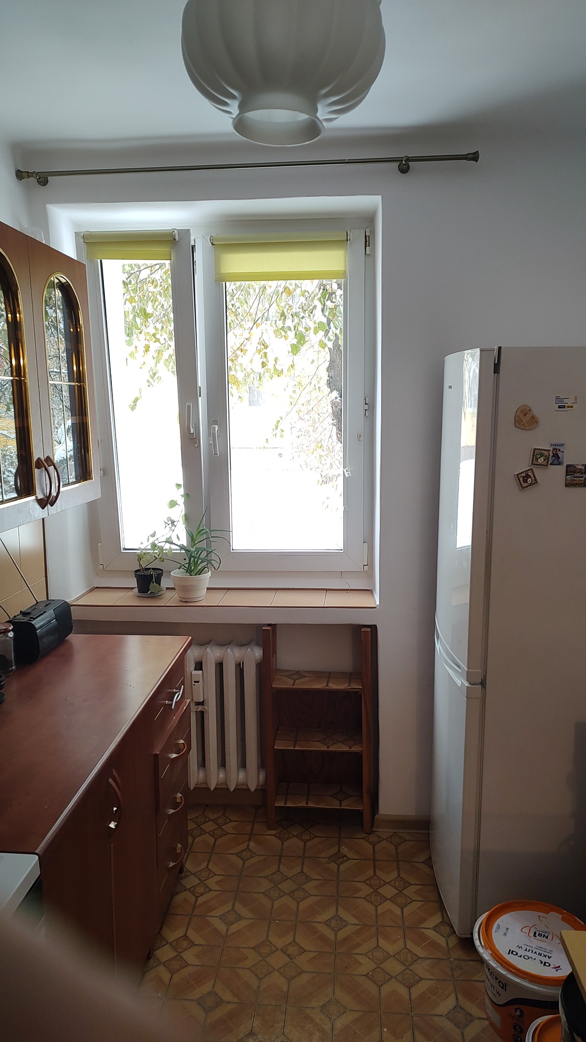 Fragment kuchni po remoncie, widoczne okno z roletami, grzejnik, szafki, lodówka z magnesami i wiadro z farbą, ukazujące etap wykończenia.