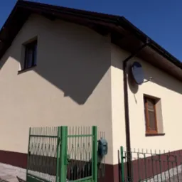 Dom jednorodzinny z jasną elewacją i ciemnobrązowym cokołem, widoczne okna z brązowymi ramami, antena satelitarna i zielone ogrodzenie.