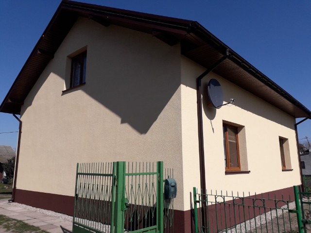 Dom jednorodzinny z jasną elewacją i ciemnobrązowym cokołem, widoczne okna z brązowymi ramami, antena satelitarna i zielone ogrodzenie.