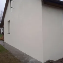 Świeżo otynkowana, jasna elewacja domu jednorodzinnego z ciemnobrązowym dachem i rynną, widoczny fragment chodnika i trawnika po deszczu.