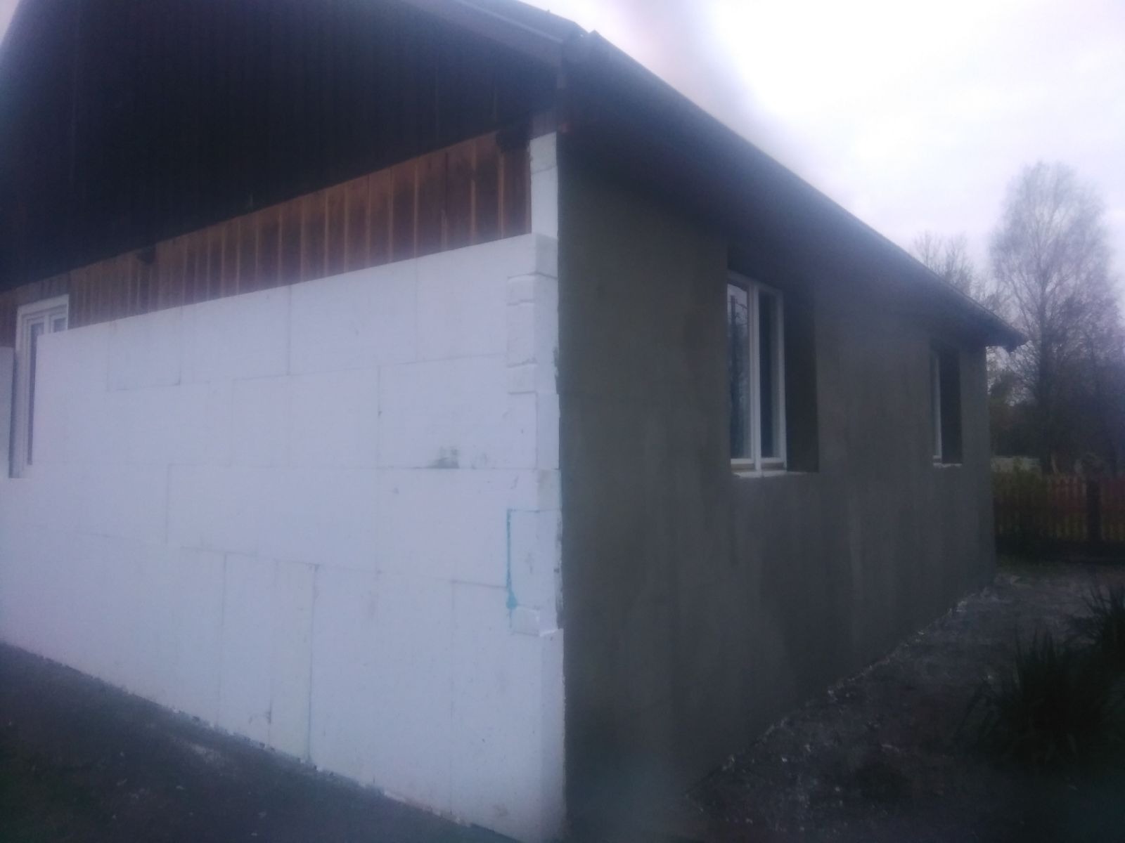 Fragment budynku w trakcie prac: jedna ściana z ociepleniem styropianowym, druga z nałożonym szarym tynkiem. Widoczne okna i drewniane elementy dachu.