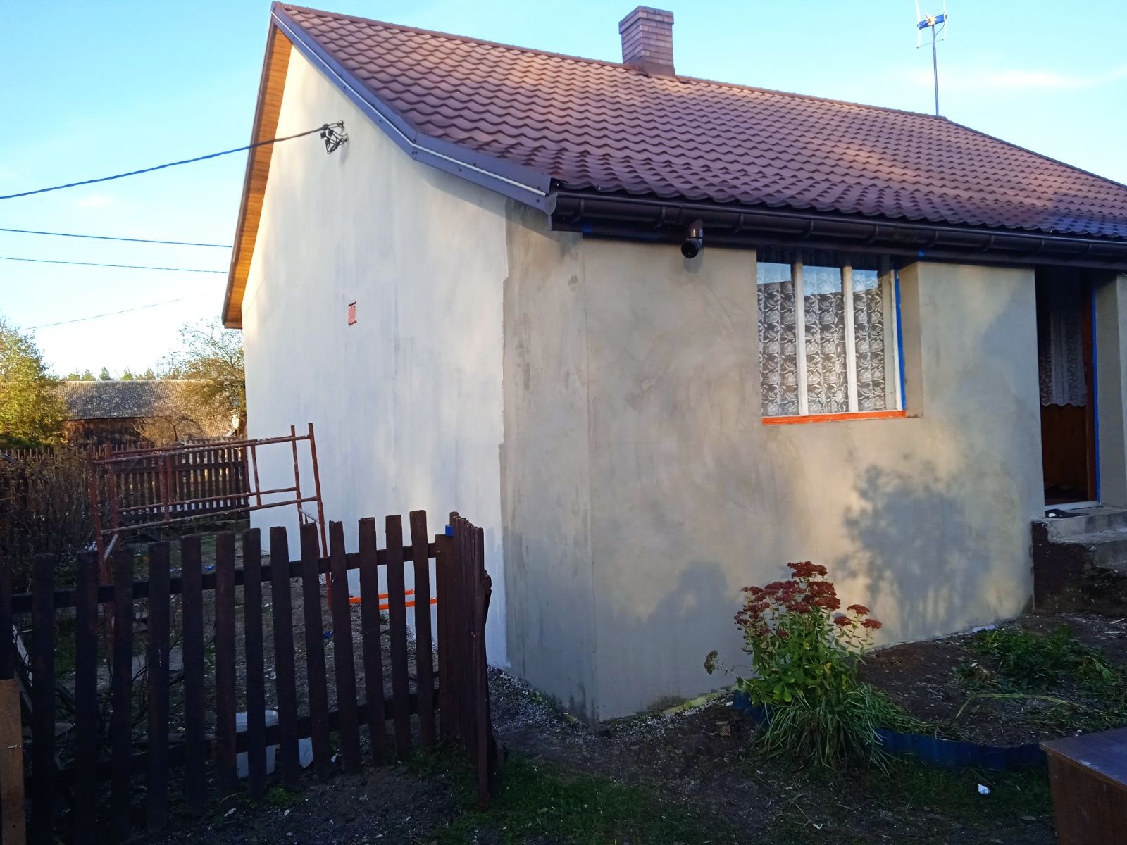 Dom jednorodzinny po nałożeniu szarej warstwy tynku przed ociepleniem, widoczne rusztowanie, okno z pomarańczową ramą zabezpieczone folią malarską i brązowy dach z ceglanym kominem.