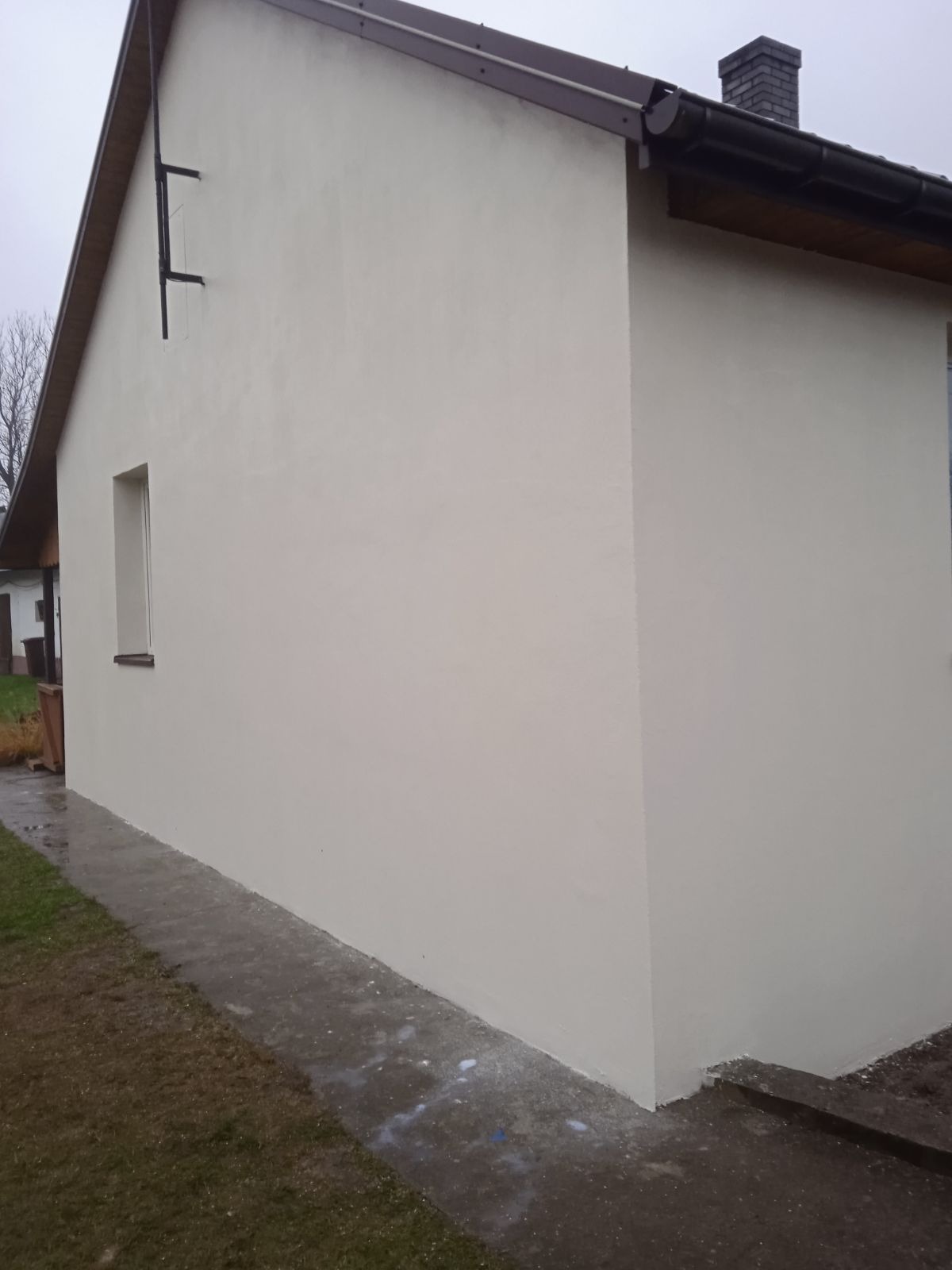 Świeżo otynkowana, jasna elewacja domu jednorodzinnego z ciemnobrązowym dachem i rynną, widoczny fragment chodnika i trawnika po deszczu.