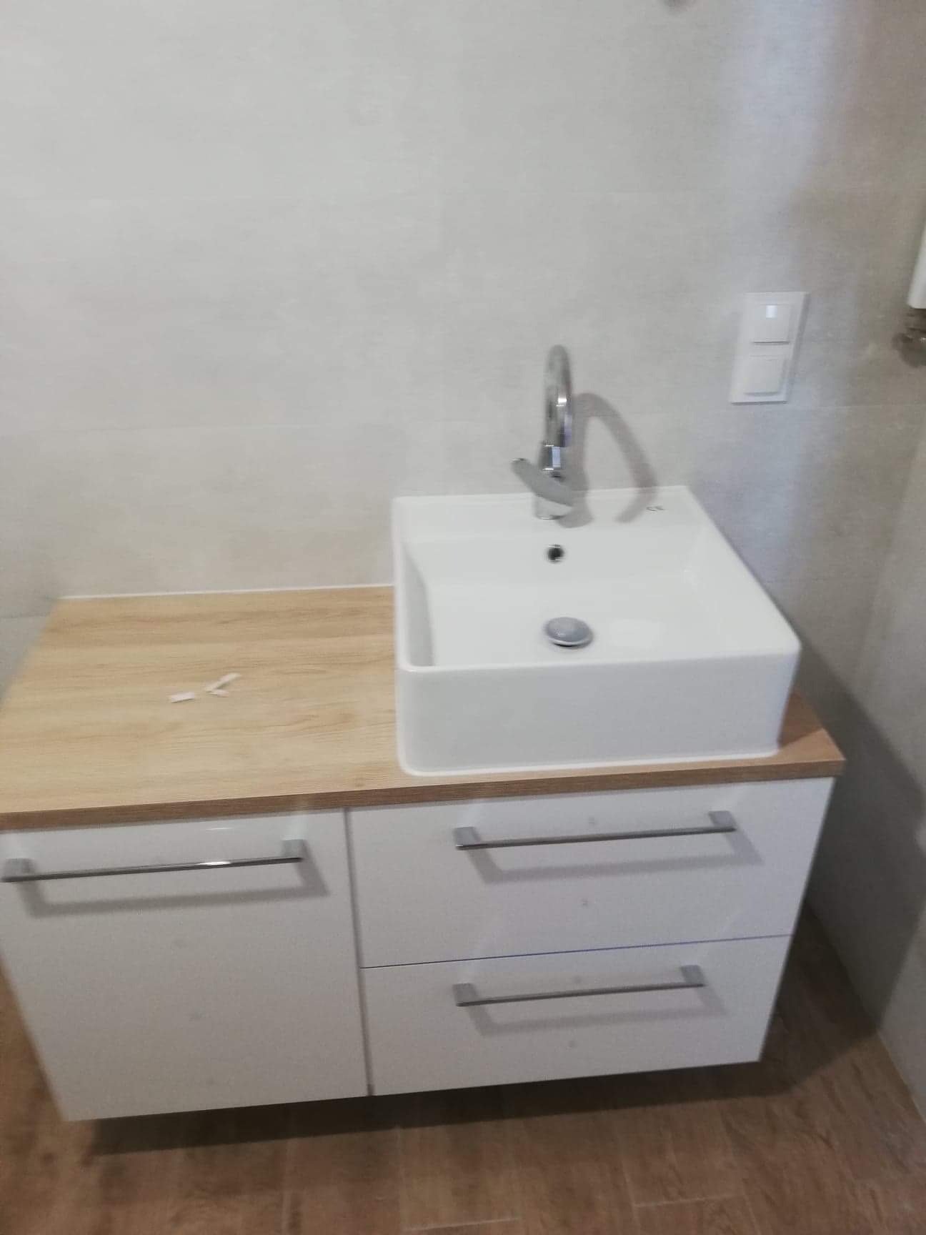 Nowoczesna, kwadratowa umywalka nablatowa z chromowaną baterią, osadzona na szafce z białymi frontami i blatem imitującym jasne drewno. Nad umywalką widoczny włącznik światła na ścianie wyłożonej...