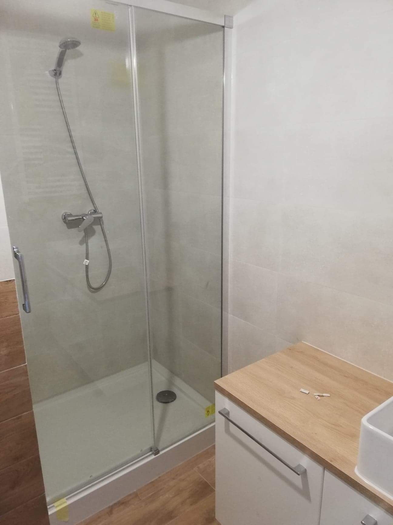 Nowoczesna kabina prysznicowa z przesuwnymi drzwiami, jasne płytki na ścianach i podłodze imitują drewno, widoczna szafka pod umywalką z blatem w kolorze jasnego drewna, fragmenty taśmy malarskiej...