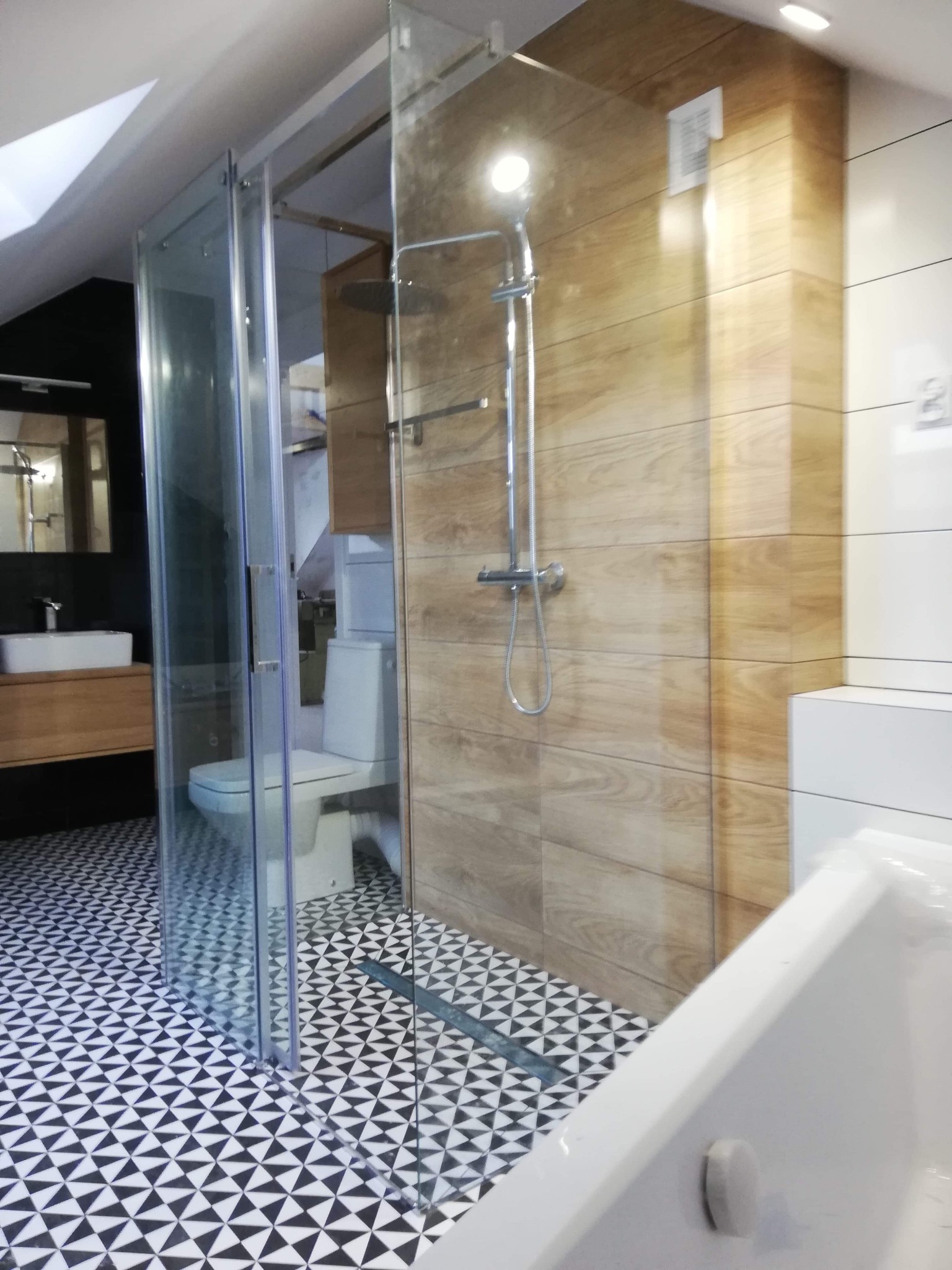 Nowoczesna łazienka na poddaszu z kabiną prysznicową typu walk-in, drewnopodobnymi płytkami ściennymi, czarno-białą mozaiką podłogową i minimalistyczną umywalką na drewnianej szafce.