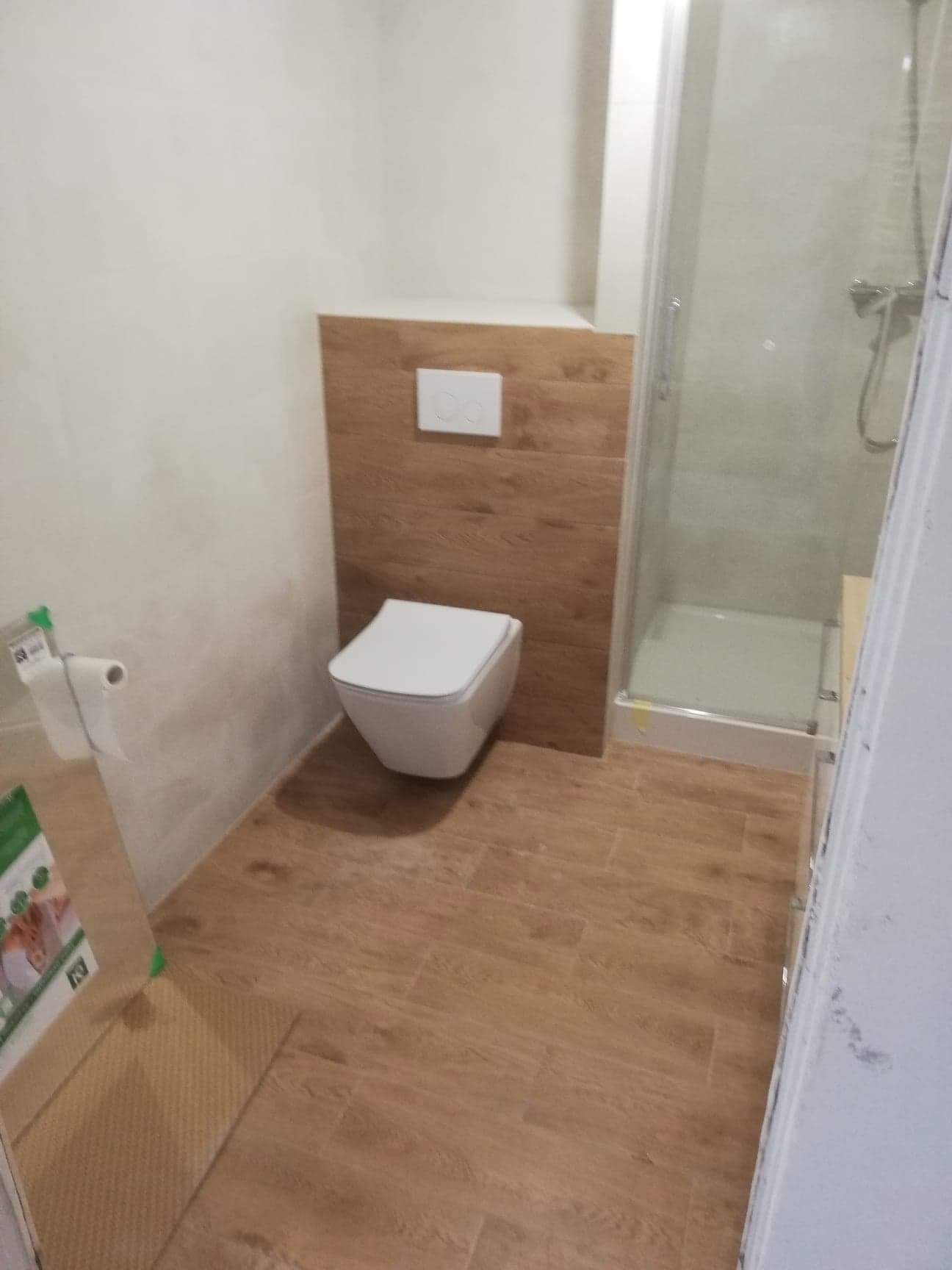 Nowoczesna łazienka z podwieszaną toaletą, płytkami imitującymi drewno na podłodze i ścianie, oraz kabiną prysznicową z przezroczystego szkła.