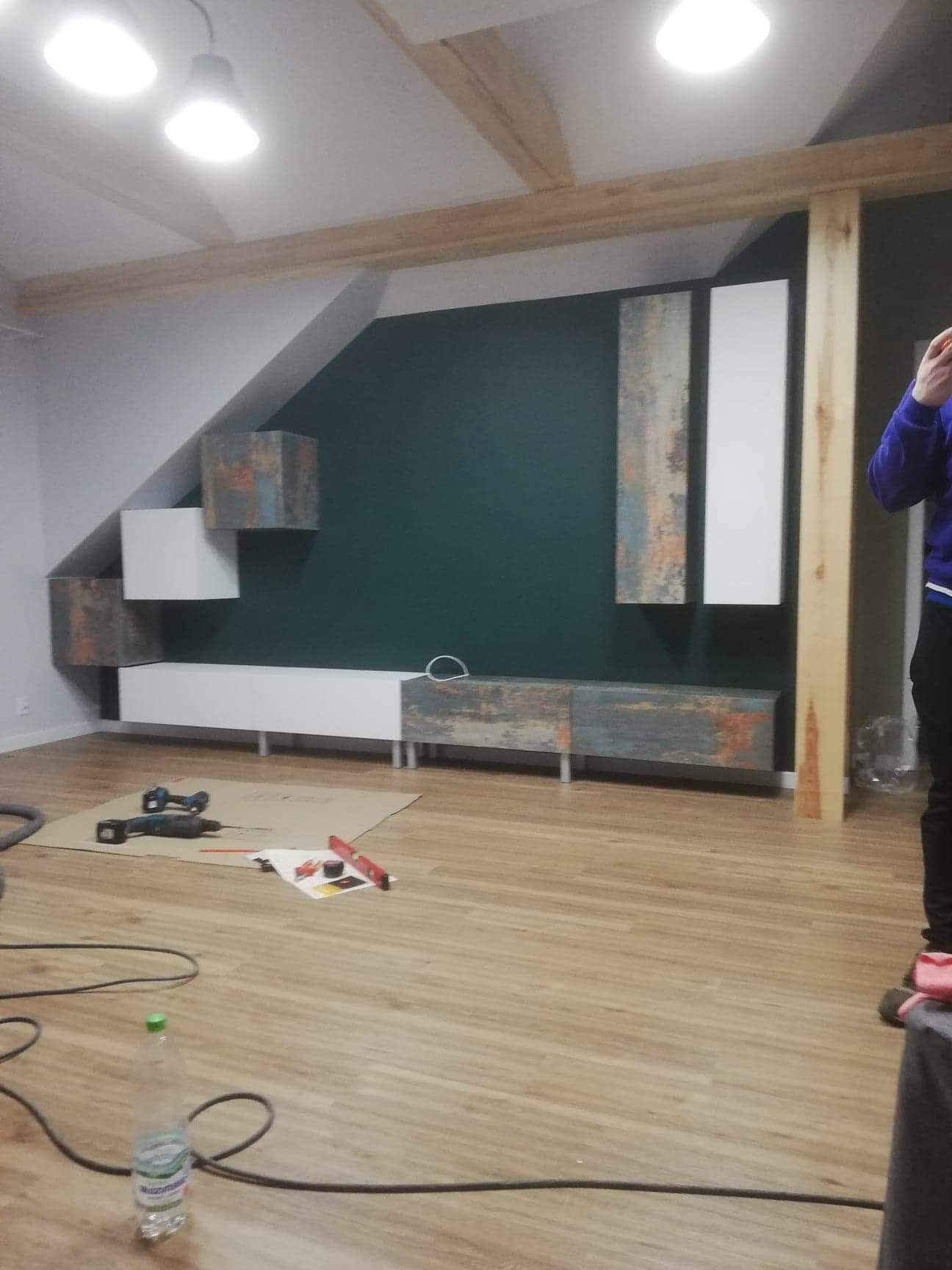 Pokój z zamontowanymi meblami na ścianie w trakcie prac wykończeniowych, widoczne narzędzia, karton ochronny i butelka wody na podłodze z paneli.