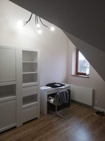 Pokój na poddaszu z drewnianą podłogą, białymi meblami, biurkiem i oknem w skosie dachu, oświetlony lampą sufitową z kilkoma żarówkami.