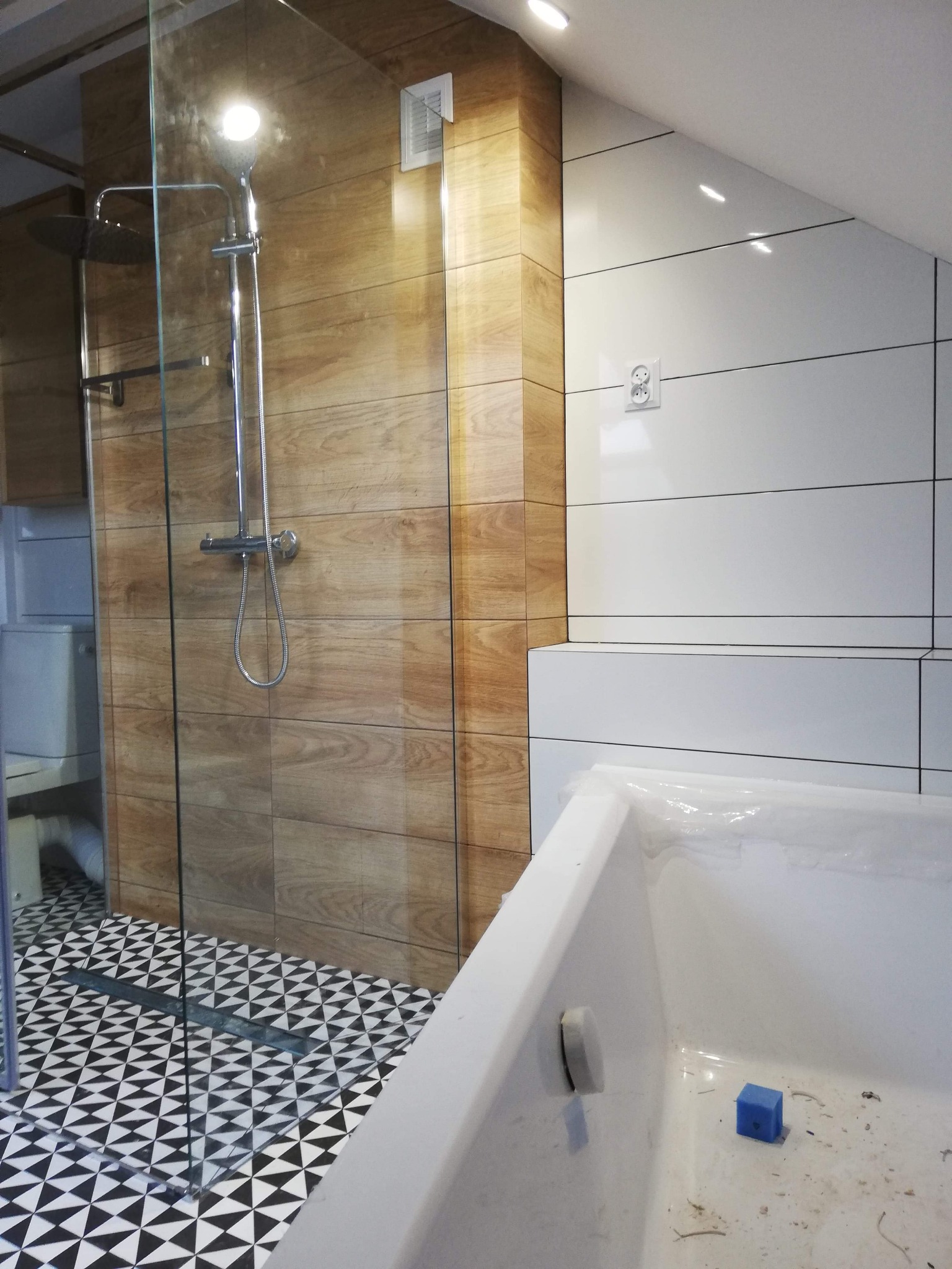 Nowoczesna łazienka na poddaszu z kabiną prysznicową typu walk-in, wykończona płytkami imitującymi drewno, białą ceramiką i geometryczną posadzką w czarno-białe trójkąty.
