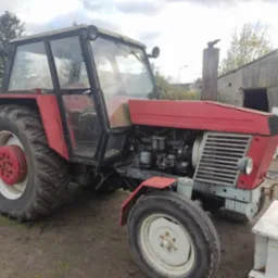 Czerwony traktor Ursus z kabiną na tle wiejskiego krajobrazu z drewnianym budynkiem gospodarczym i stertą gałęzi.