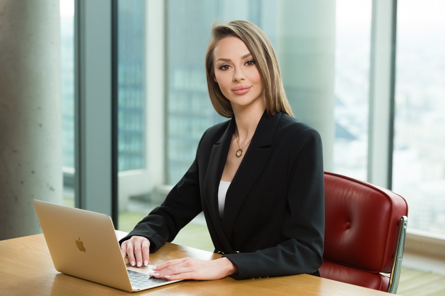 Profesjonalny portret kobiety w czarnym żakiecie pracującej na laptopie w nowoczesnym biurze z widokiem na miasto.