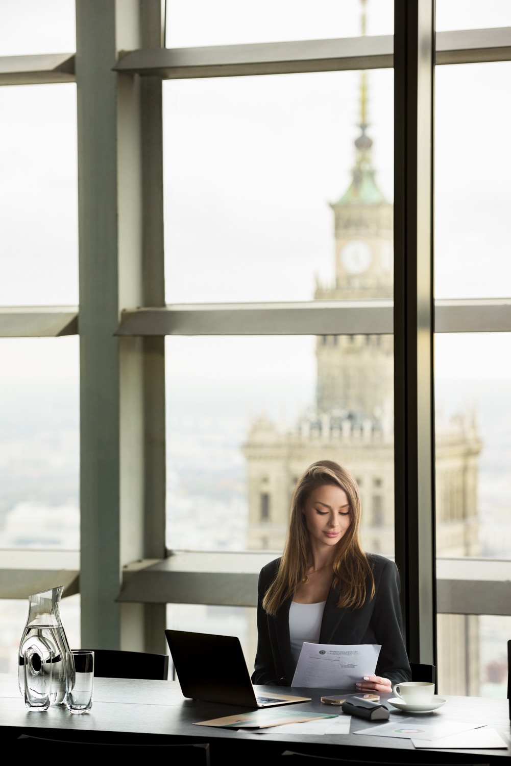 Kobieta w biurze czyta dokumenty, za oknem widok na Pałac Kultury w Warszawie, na biurku laptop, woda w karafce i filiżanka kawy.