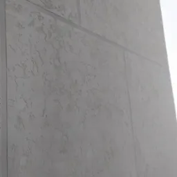 Fragment szarej elewacji z wyraźną fakturą, podzielonej na panele z widocznymi łączeniami, oświetlonej z prawej strony.