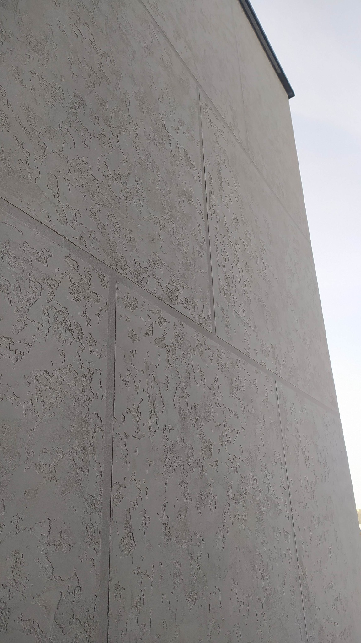 Fragment ściany budynku z szarą elewacją o wyraźnej fakturze, podzielonej na panele pionowymi i poziomymi liniami, widzianej z dołu pod kątem.