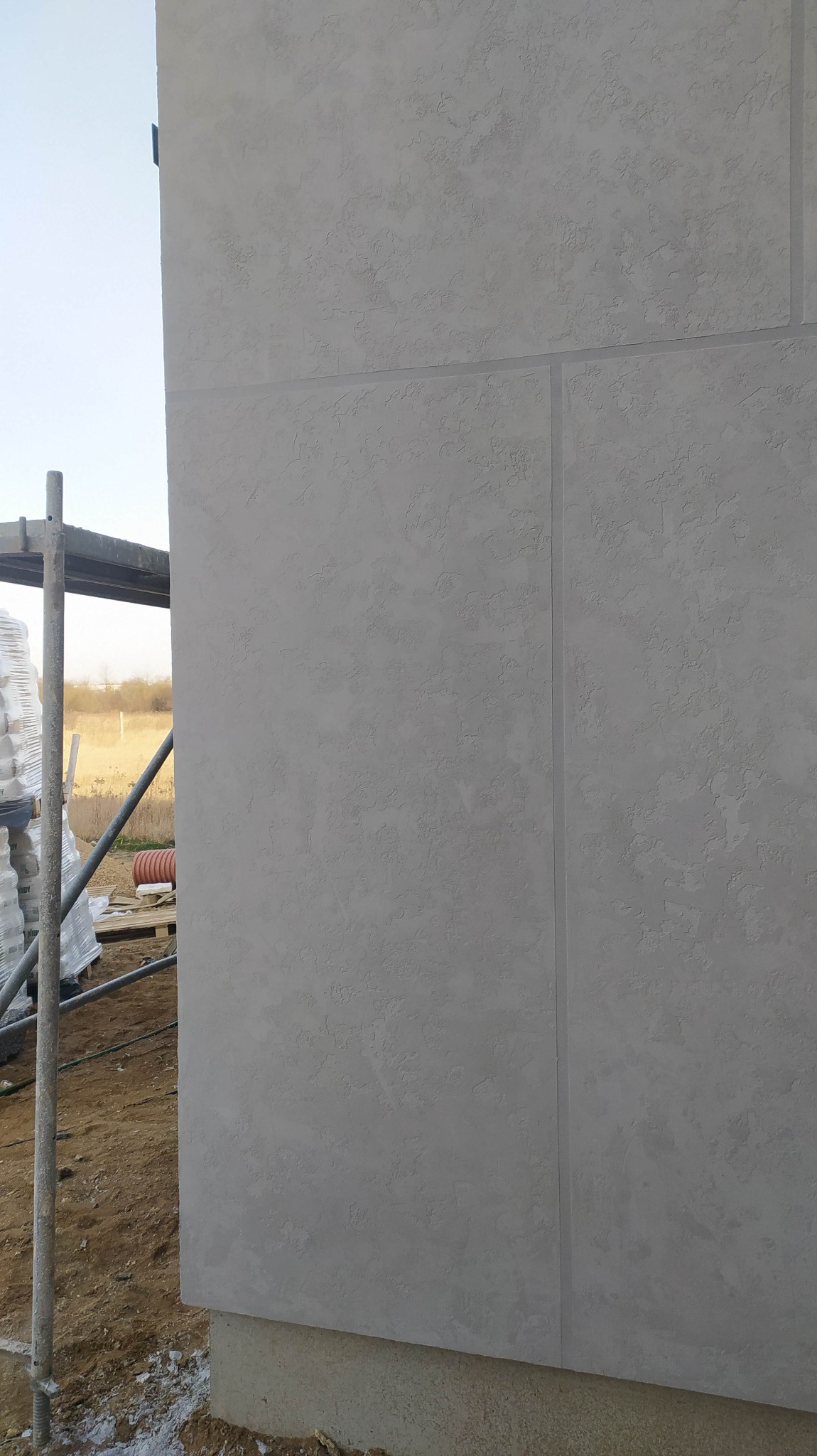 Fragment elewacji budynku z dekoracyjnym tynkiem mineralnym w odcieniach szarości, widoczne łączenia płyt elewacyjnych oraz rusztowanie z workami materiałów budowlanych w tle.