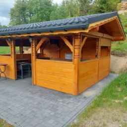 Drewniana konstrukcja z zadaszeniem krytym dachówką, z elementami dekoracyjnymi, na wybrukowanym podłożu, z grillem widocznym wewnątrz.