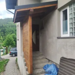 Drewniana wiata nad wejściem do domu z widocznymi schodami, elewacją i oknem z luksferami, w tle zalesione wzgórza.