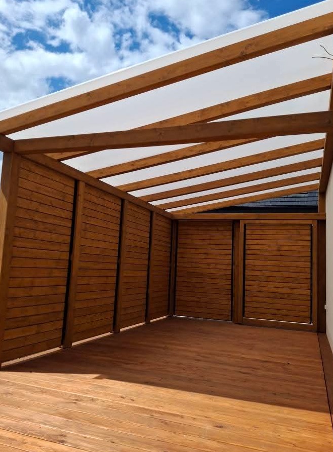 Zadaszona, drewniana konstrukcja z deskami w odcieniach brązu, tworząca zacienione miejsce z drewnianą podłogą. Widoczne niebo z chmurami w tle.