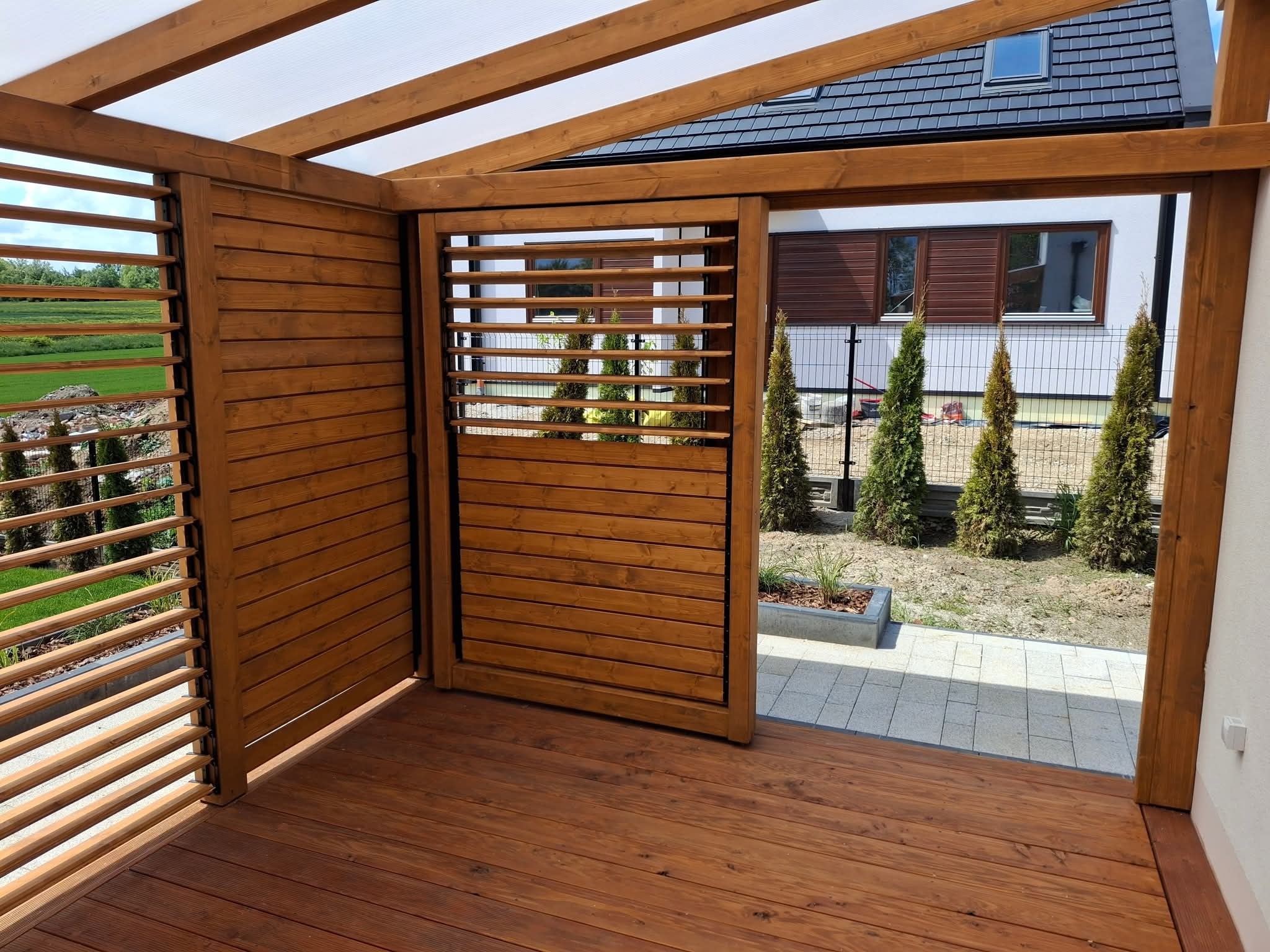 Drewniana pergola z przesuwnymi panelami lamelowymi, widok z wnętrza na ogród i dom. Ciemne drewno, jasne zadaszenie, nowoczesny design.