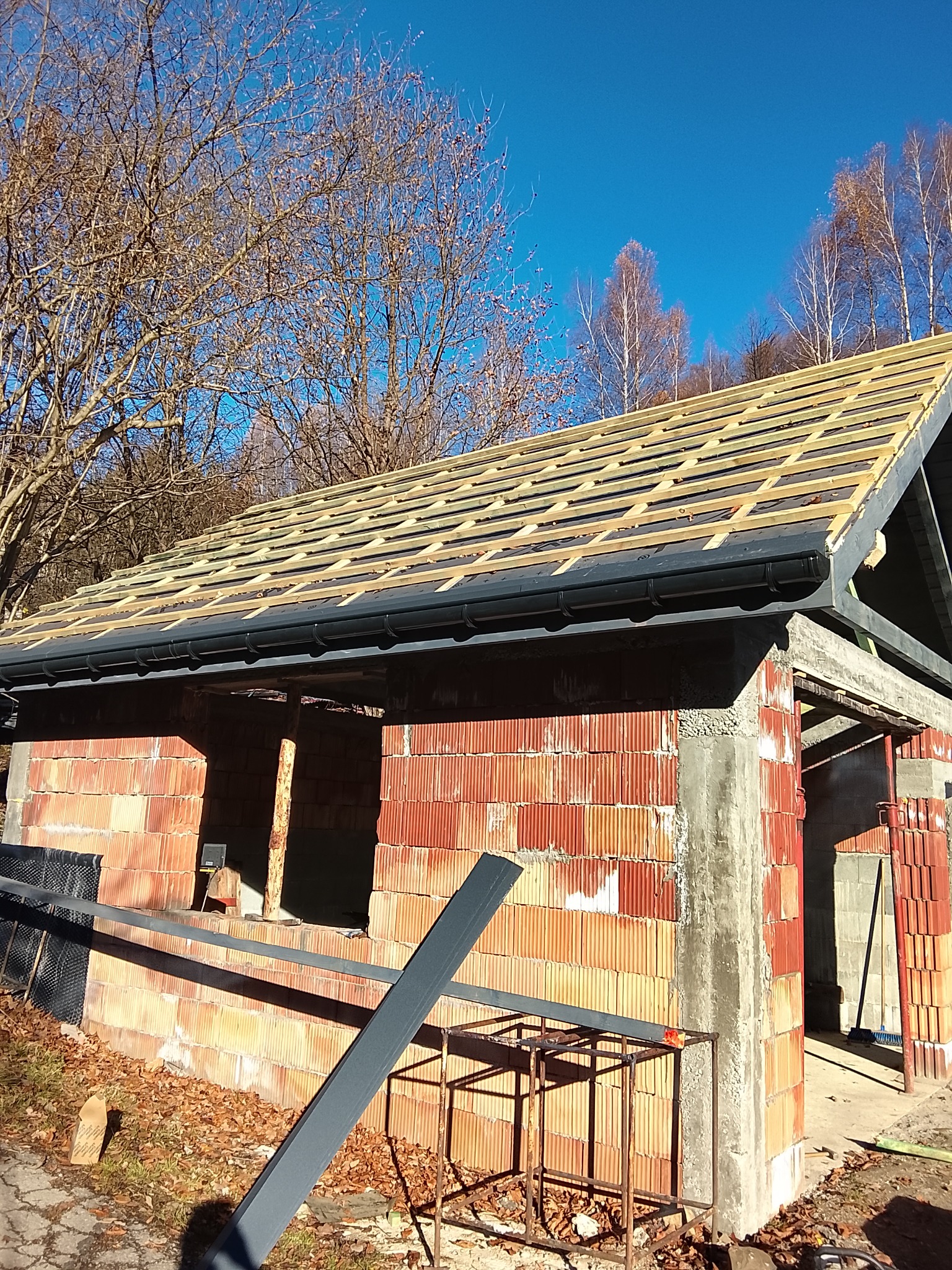 Wznoszona konstrukcja dachu z drewnianą łatą i czarnymi rynnami, na tle ceglanego budynku w stanie surowym otwartym i błękitnego nieba.