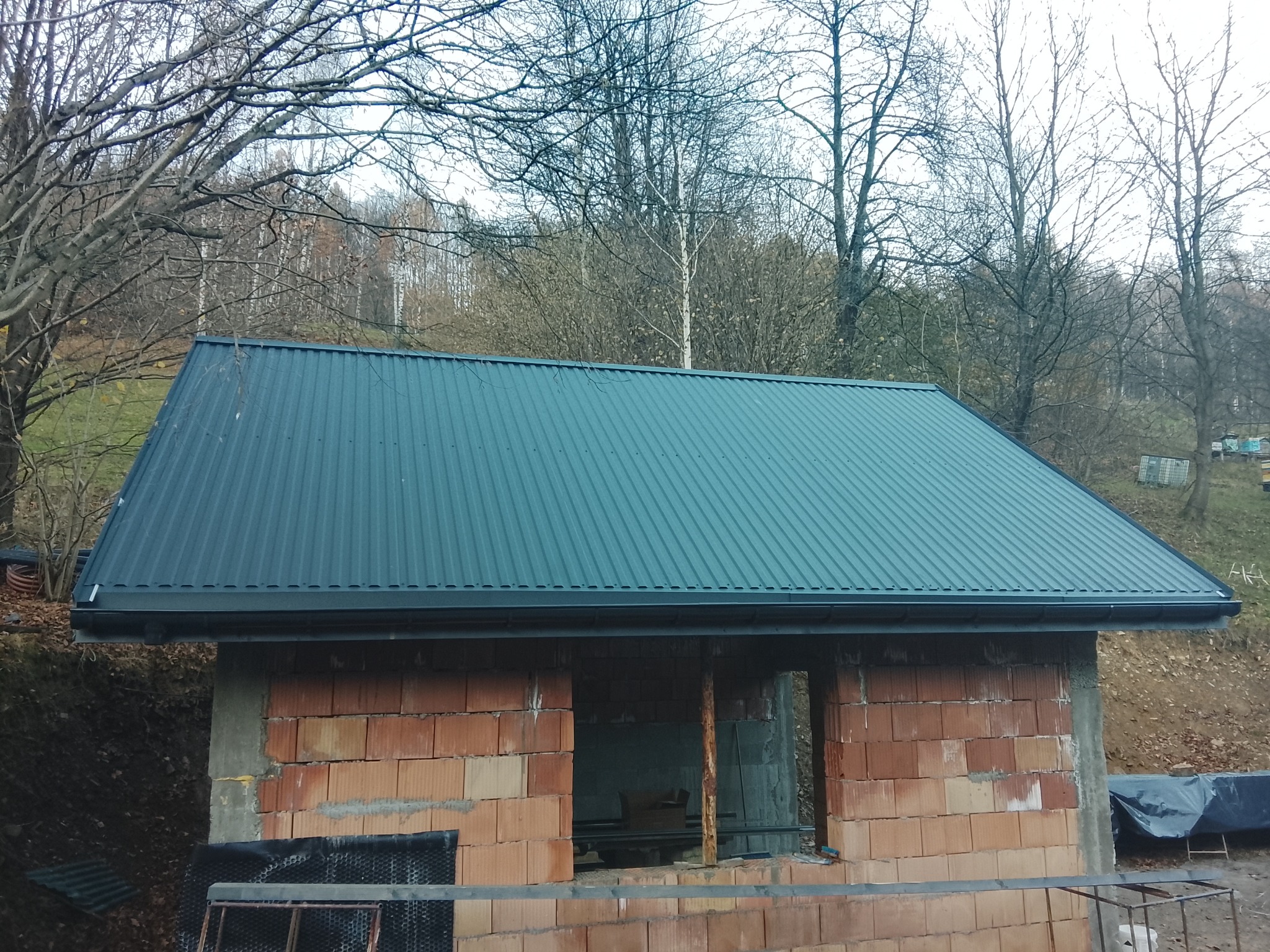 Niebieski dach z blachy trapezowej na budynku w trakcie budowy, widoczne cegły i rynny. W tle drzewa bez liści i pagórkowaty teren.