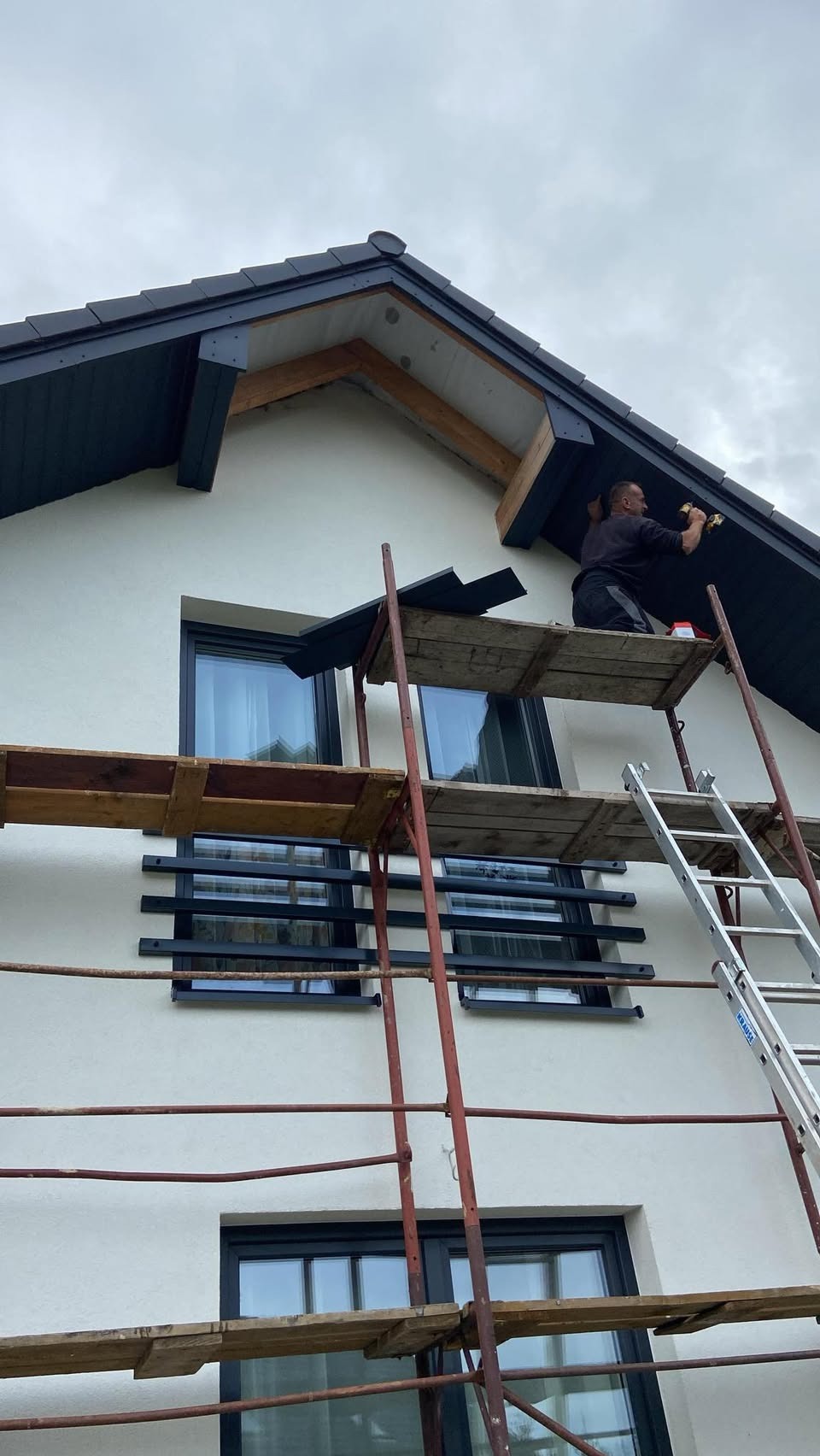 Pracownik na rusztowaniu montuje podbitkę dachową w nowoczesnym domu z białymi ścianami i czarnymi oknami. Widoczne narzędzia i fragmenty podbitki.