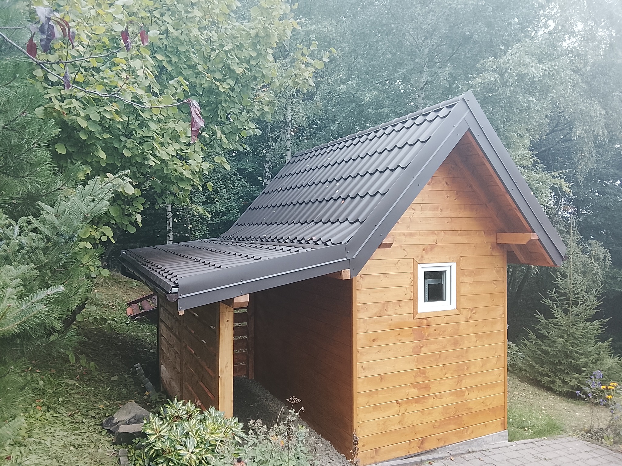 Drewniana altana z ciemnym dachem w Szczyrku, otoczona zielenią. Widoczne małe okno i konstrukcja wsparta na słupach, tworząca zadaszone miejsce.