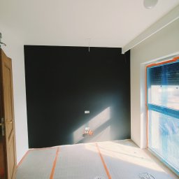 Usługi Remontowo Budowlane Mateusz Bartoszek - Pokój w trakcie prac, z czarną ścianą, zabezpieczoną folią okienną i podłogą wyłożoną folią ochronną. Widoczne puszki z farbą i narzędzia malarskie.