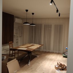 Usługi Remontowo Budowlane Mateusz Bartoszek - Jasne wnętrze z drewnianą podłogą i stołem. Widoczne oświetlenie sufitowe, firany w oknach i elementy wykończenia.