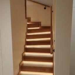 Usługi Remontowi Budowlane Mateusz Bartoszek - Drewniane schody z podświetleniem LED, widok z dołu. Naturalne drewno, minimalistyczny design, ciepłe światło podkreśla strukturę drewna.