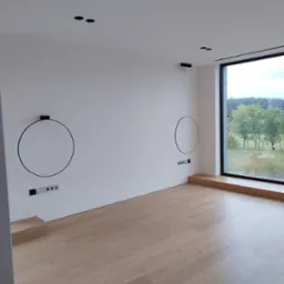 Minimalistyczne wnętrze z drewnianą podłogą, białymi ścianami i dużym oknem z widokiem na zieleń. Dekoracyjne, okrągłe lampy ścienne.