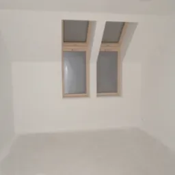 Poddasze z dwoma oknami dachowymi w drewnianych ramach, ściany i sufit pomalowane na biało, widoczne ślady po pracach wykończeniowych na podłodze.