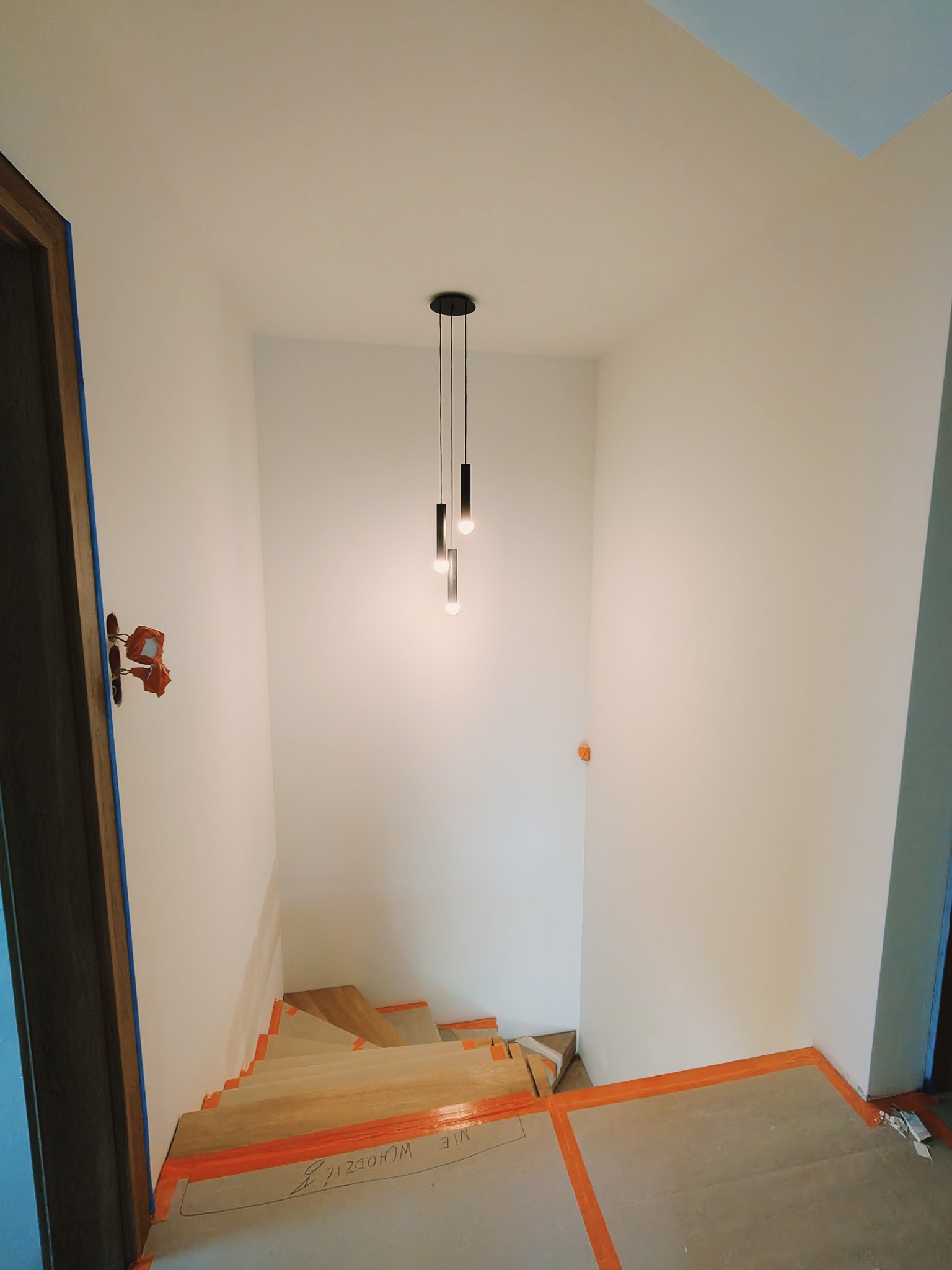 Wnętrze klatki schodowej w trakcie remontu, zabezpieczone folią i taśmą malarską, z widocznymi schodami w surowym drewnie i nowoczesną lampą wiszącą.