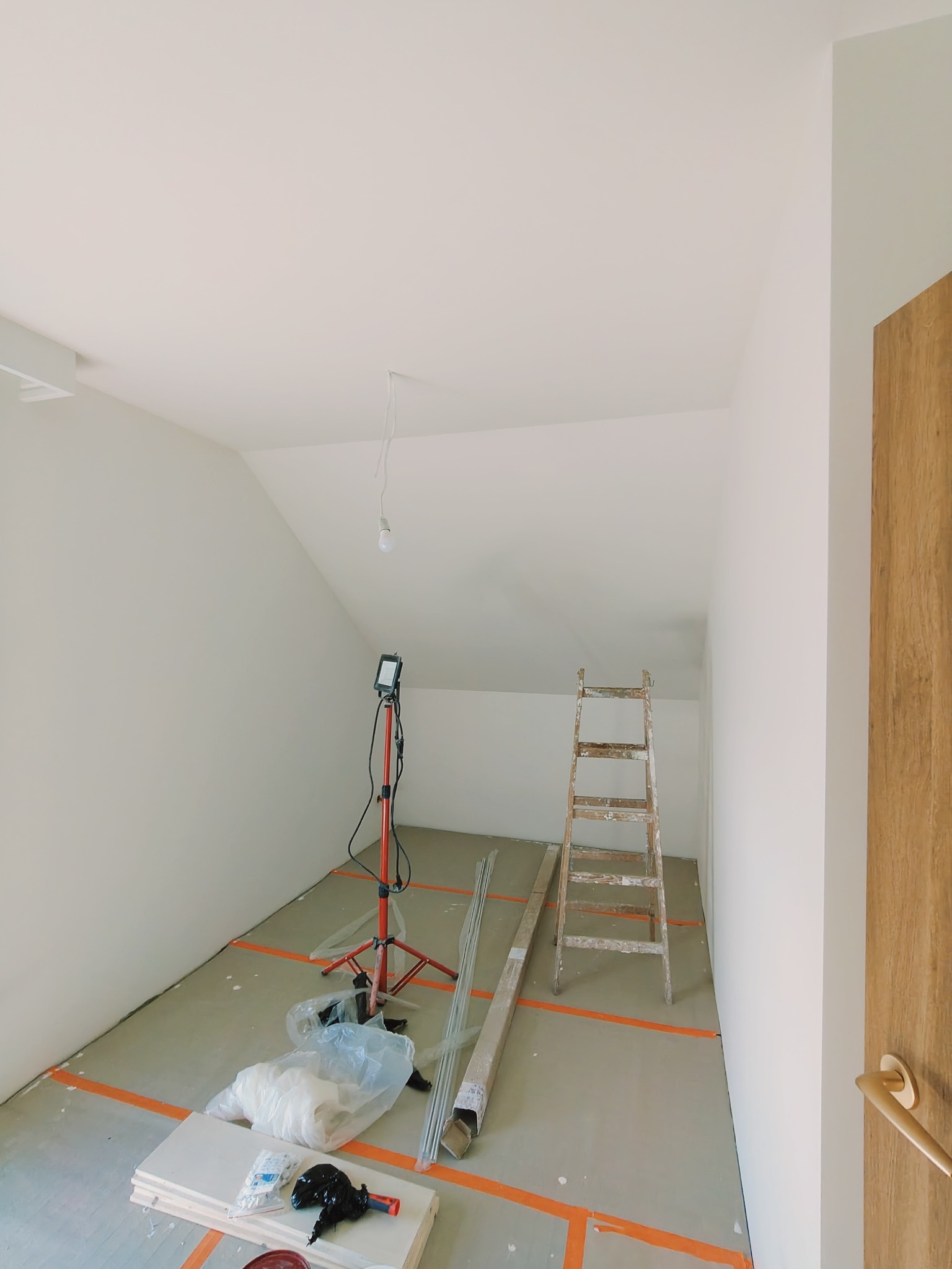 Poddasze w trakcie prac wykończeniowych: widoczna drabina, lampa oświetleniowa na statywie, profile, płyty i narzędzia zabezpieczone folią na podłodze, pomalowane ściany i sufit na biało,...