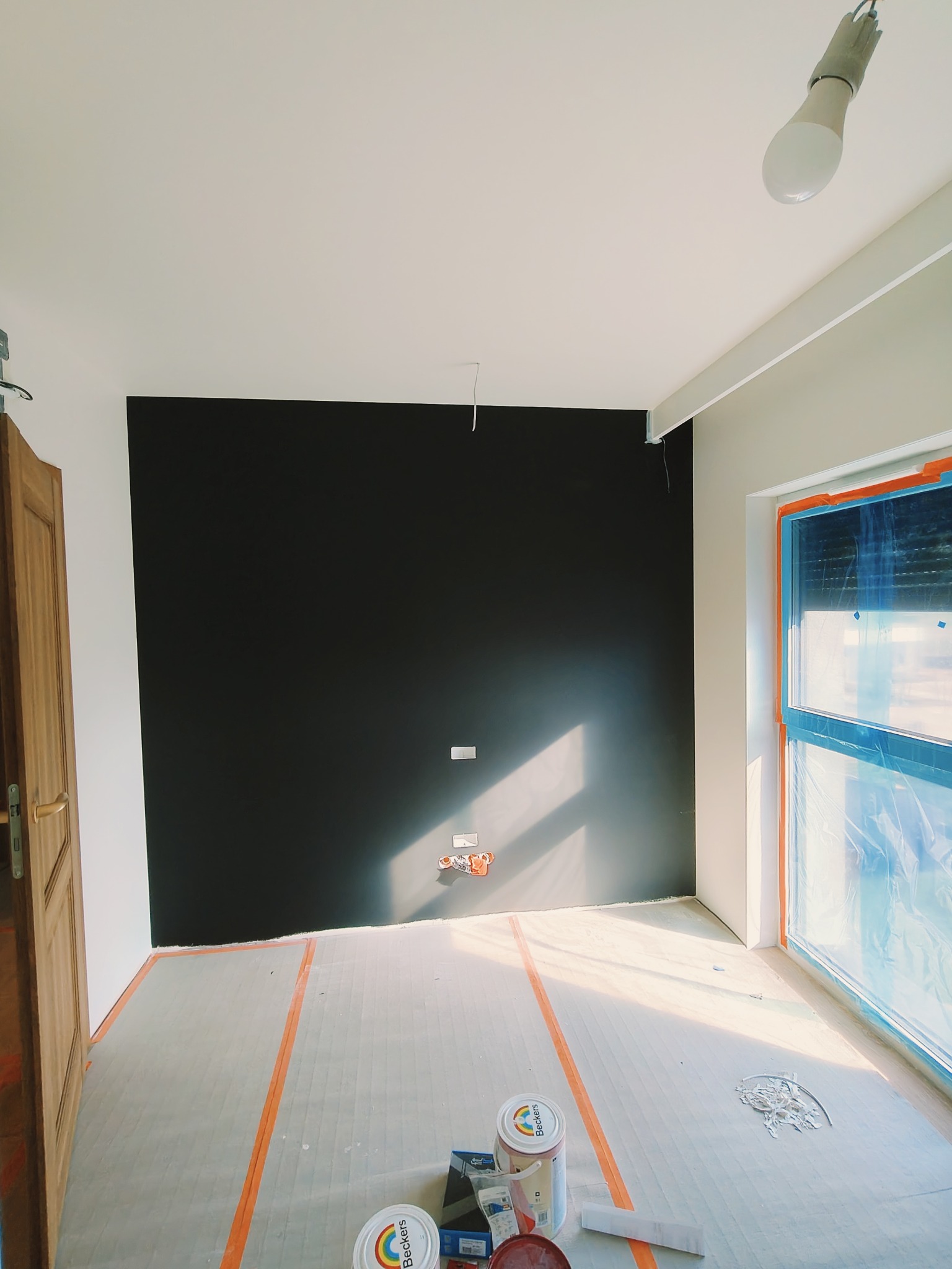 Pokój w trakcie prac, z czarną ścianą, zabezpieczoną folią okienną i podłogą wyłożoną folią ochronną. Widoczne puszki z farbą i narzędzia malarskie.